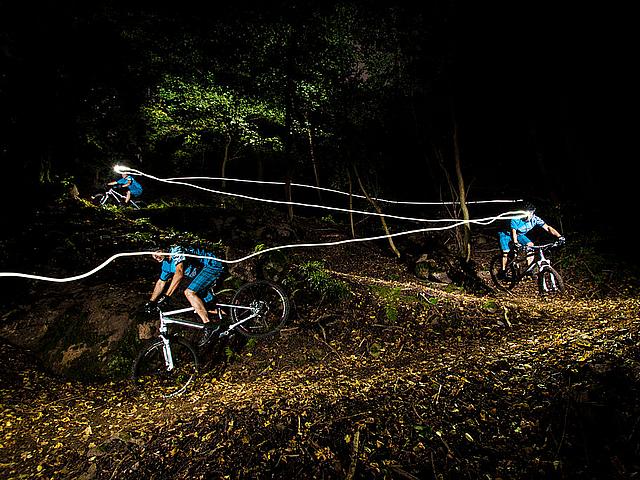 drei Fahrradfahrer bei Nacht auf einem Waldweg, Kopflampen hinterlassen Lichtspur