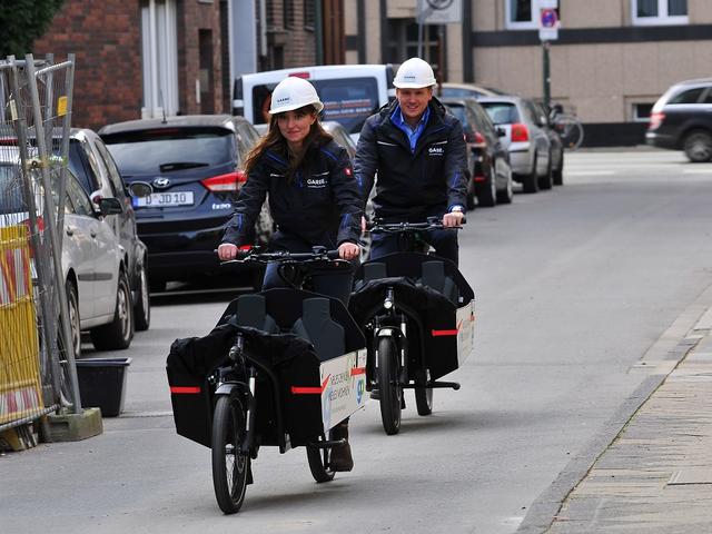 Vorne eine Frau, dahinter ein Mann. Beide mit weißem Helm auf schwarzen Lastenrädern.