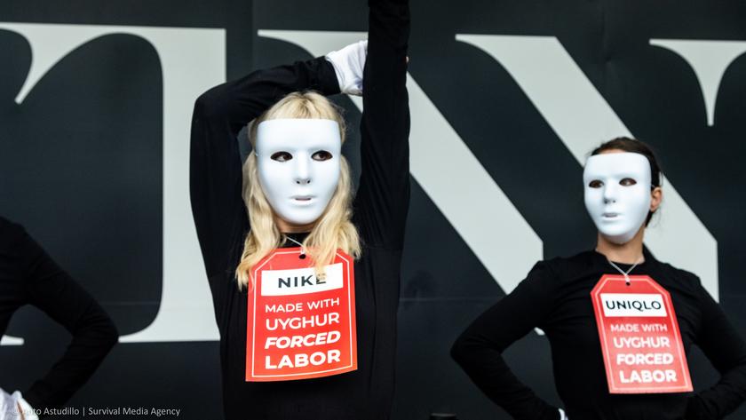 Zwei Menschen hinter weißen Masken tragen Schilder auf denen steht "Nike/Uniqlo made with Uyghur forced Labour"