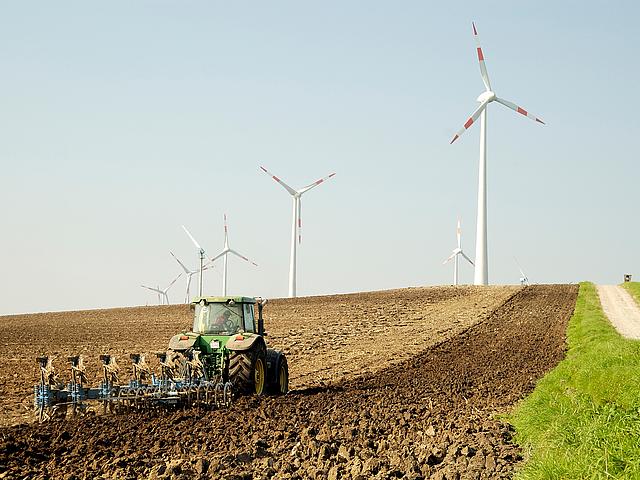Ein Traktor auf einem Feld. Dahinter ragen Windkraftanlagen in die Höhe.