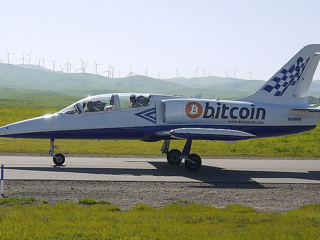 Foto: Bild eines Düsenjets mit der Aufschrift Bitcoin. Im Hintergrund Windräder.