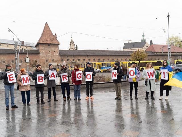 Mehrere Personen halten auf einem Platz Buchstaben hoch, die zusammen "Embargo Now" ergeben. Zudem hält eine Frau eine Flagge der Ukraine. 