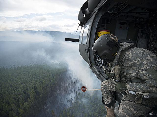 Ein Soldat in Uniform sitzt in einem Helikopter und steuert den Abwurf von Wasser über einem Waldbrand. 