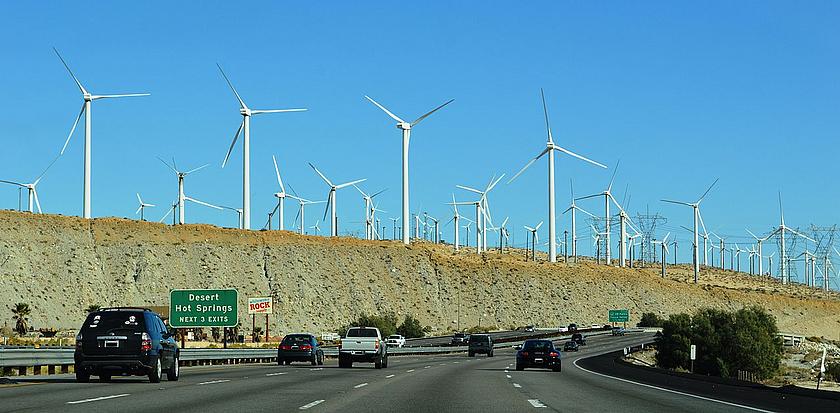 Dutzende Windräder in einer Wüstenlandschaft ragen über einer vielspurigen Autobahn auf.