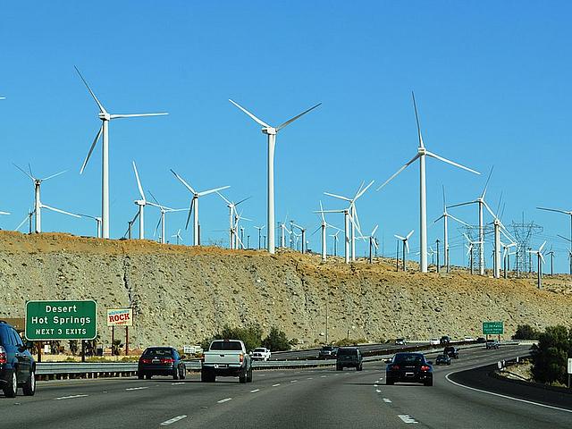 Dutzende Windräder in einer Wüstenlandschaft ragen über einer vielspurigen Autobahn auf.