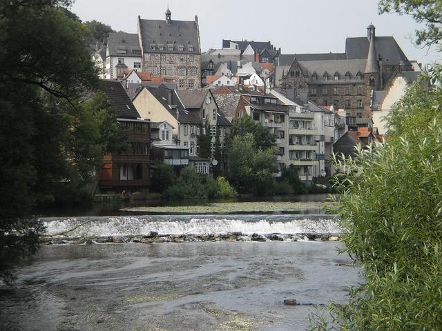 Blick auf den Fluss Lahn mit einem Wehr, im Hintergrund die Stadt Marburg