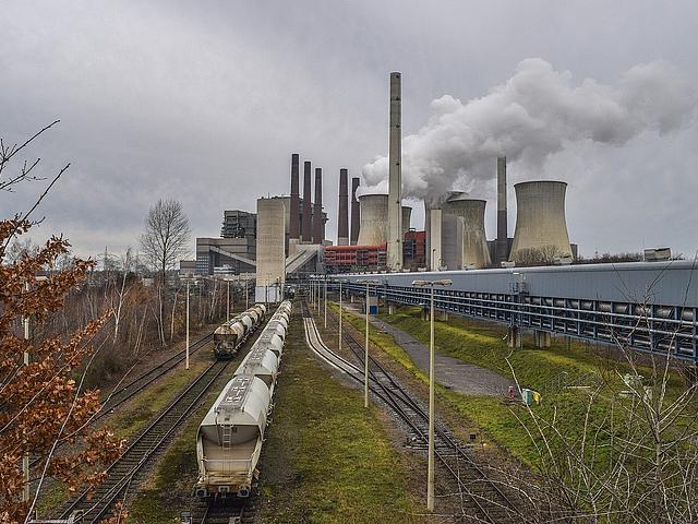Ein Kohlekraftwerk im Hintergrund, mit Kohletransportern auf Schienen im Vordergrund.