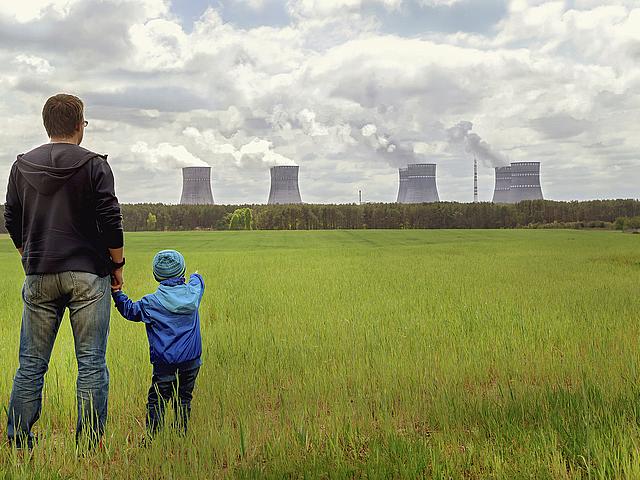 Vater steht mit seinem Kind auf einem Feld. Vor ihnen steht ein Kohlekraftwerk mit rauchenden Schloten.