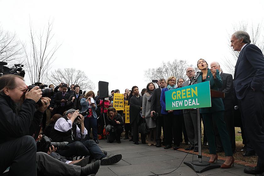 Bild von Alexandria Ocasio-Cortez, hinter einem Rednerpult mit der Aufschrift Green New Deal. Davor viele Kameraleute.