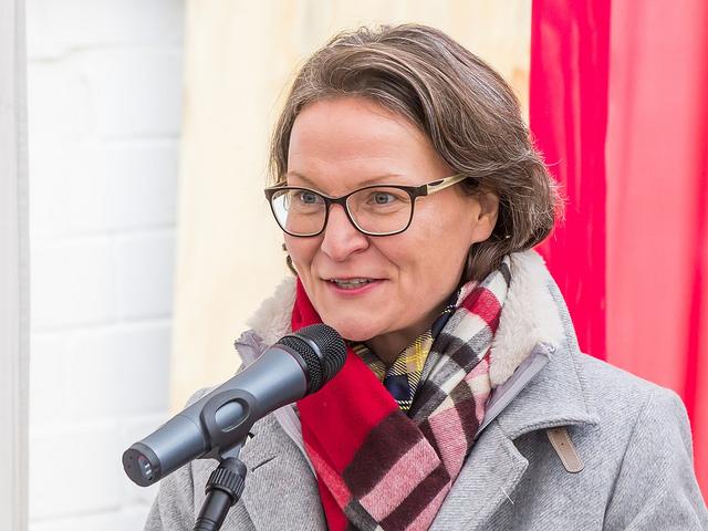 Eine Frau im grauen Mantel, rotem Schal und Brill hinter einem Mikrofon.