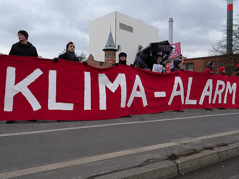 Demosntranten mit einem roten Plakat vor einem fossilen Kraftwerk in Berlin. Auf dem Plakat steht "Klima-Alarm".