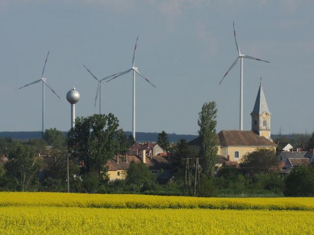 Ein Dorf mit einer Kirche. Dahinter ragen Windräder auf 