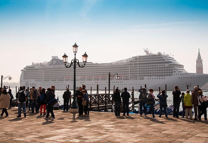 Riesiges Kreuzfahrtschiff legt an Hafen an. Im Vordergrund Menschen an einem Steg in Venedig.