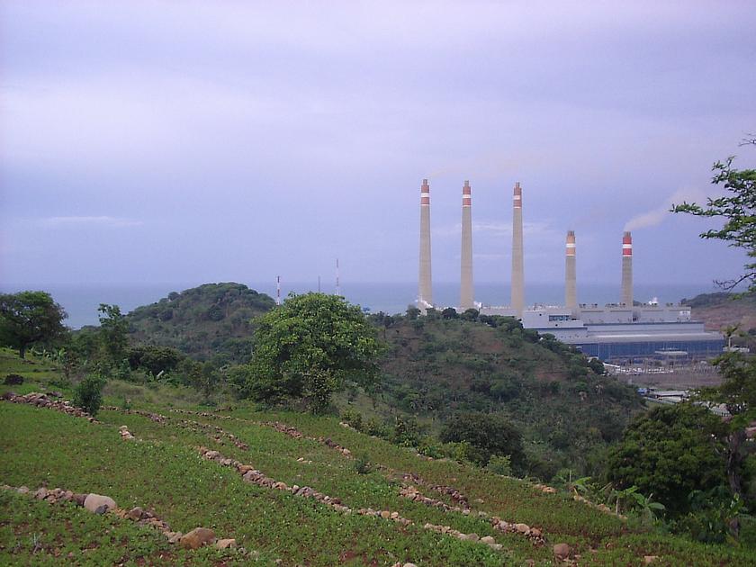 Hinter einem grünen Hügel ist ein Kohlekraftwerk am Meer zu sehen