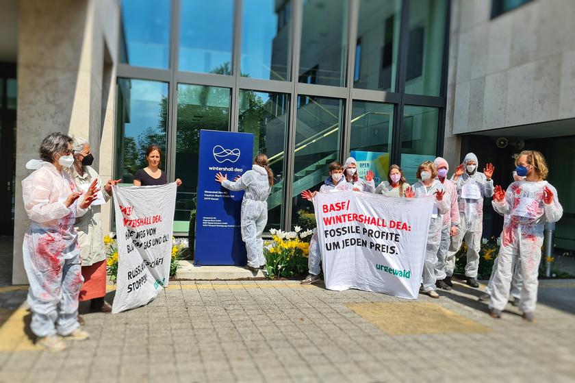 Menschen in blutbefleckten Maleranzügen vor einem Gebäudeeingang, mit Transparenten in der Hand, auf denen steht: "BASF/Wintershall Dea: Profite um jeden Preis"