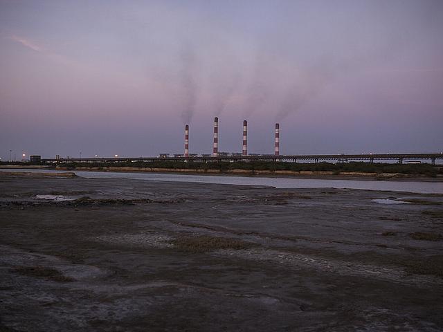 Bild eines Kraftwerks im Sonnenuntergang im Hintergrund. Im Vordergrund verbrannte Erde.