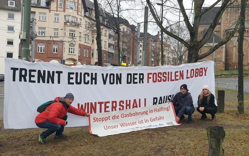 drei Menschen knien vor zwei Plakaten, auf denen steht: 1. "Trennt euch von der fossilen Lobby, Wintershall raus" 2. Stoppt die Gasbohrung in Halfing, unser Wasser ist in Gefahr.