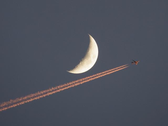 Flugzeug mit Kondensstreifen am Himmel vor beleuchtetem Planeten als Halbmond
