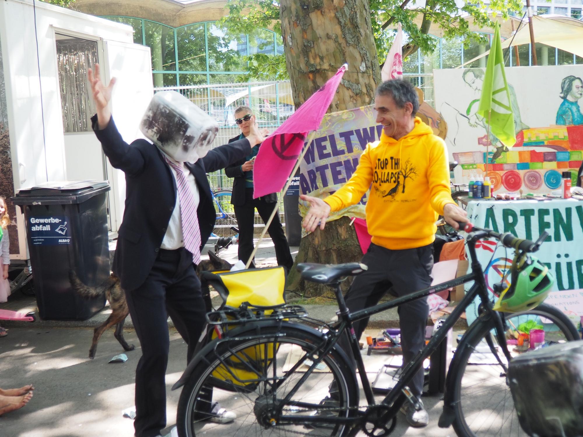 Ein Mensch im Anzug mit einem Benzinkanister über dem Kopf. Daneben ein Mann mit gelben Pullover und Fahrrad