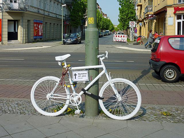 Ein weiß angemaltes Fahrrad, angeschlossen an einen Laternenpfahl, an einer Straße.