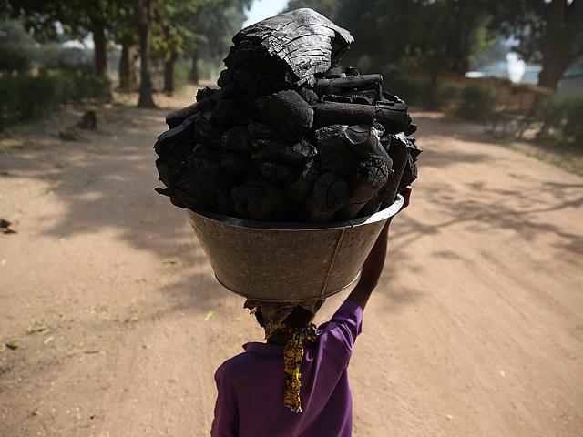 Von hinten zu sehen ist eine Frau, die einen riesigen Korb mit Kohle auf dem Kopf trägt.