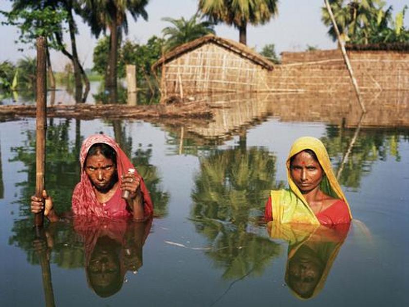 Zwei indische Frauen bis zu den Schultern im Wasser stehend. Im Hintergrund Häuser und Bäume unter Wasser. 
