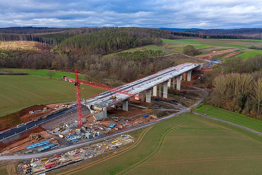 Luftbild einer im Bau befindlichen Autobahnbrücke, unweit von dichten Wäldern.