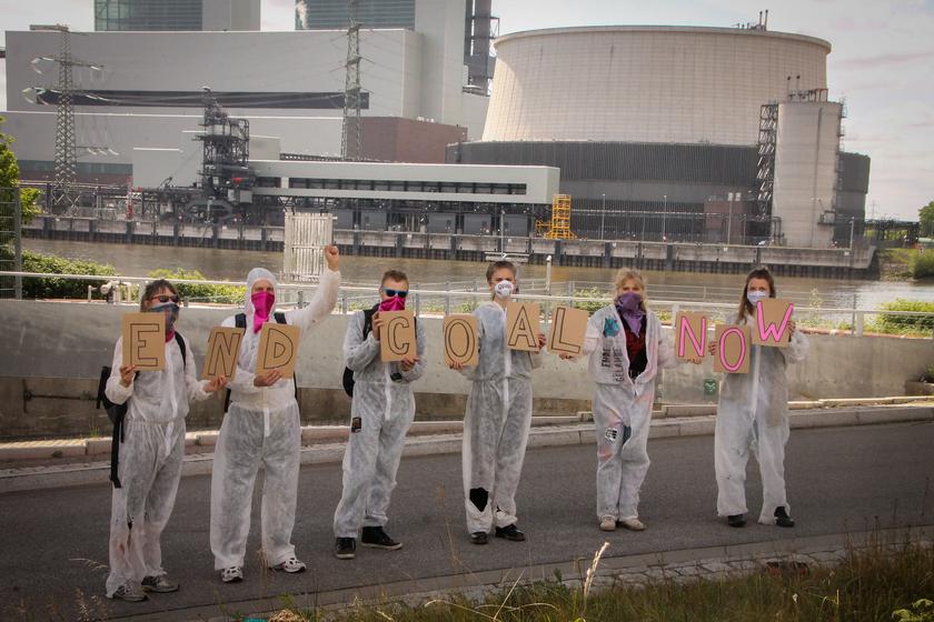 Sechs vermummte Klimaaktivist:innen in weißen Maleranzügenhalten vor einem Kraftwerk Buchstaben hoch, die zusammen "End Coal Now" ergeben 