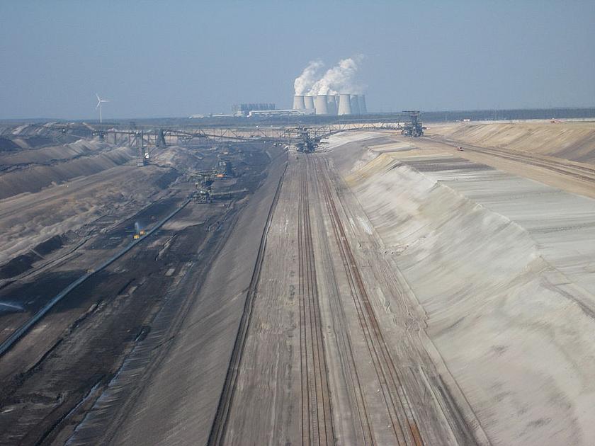 Blick auf einen Braunkohletagebau auf mehreren Ebenen. Im Hintergrund ein Kohlekraftwerk und ein Windrad. 