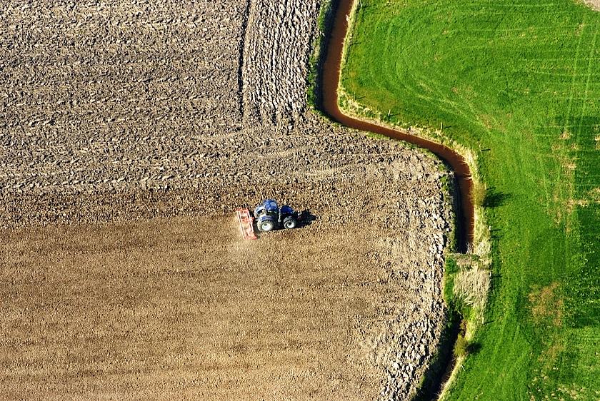 Brauner Acker mit Traktor und daneben grüne Wiese, aus der Luft fotografiert 
