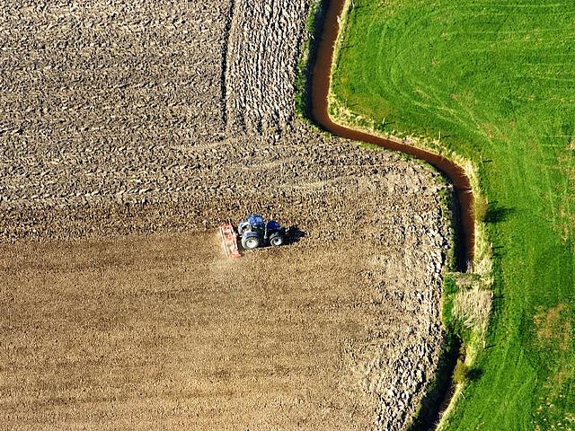 Brauner Acker mit Traktor und daneben grüne Wiese, aus der Luft fotografiert 