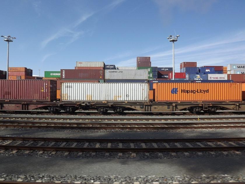 Ein Güterwaggon an einem Hafen, beladen mit Containern. Im Hintergrund stapeln sich weitere Container.