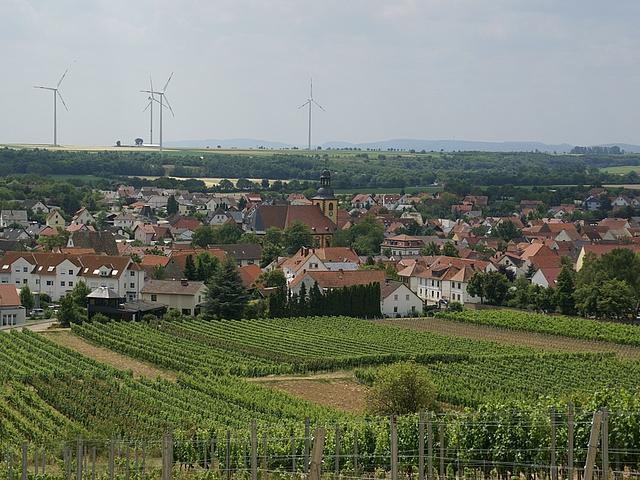 Windenergieanlagen überragen im Hintergrund ein Dorf mit einer Kirche in der Dorfmitte.