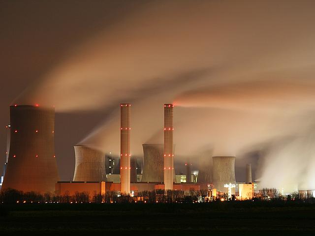 Das Kraftwerk Niederaußem bei Nacht in Langzeitbelichtung. Von Kühltürmen und Schornsteinen steigt schleierartig Wasserdampf und Rauch auf. Die Szenerie ist in gelbliches Licht getaucht.