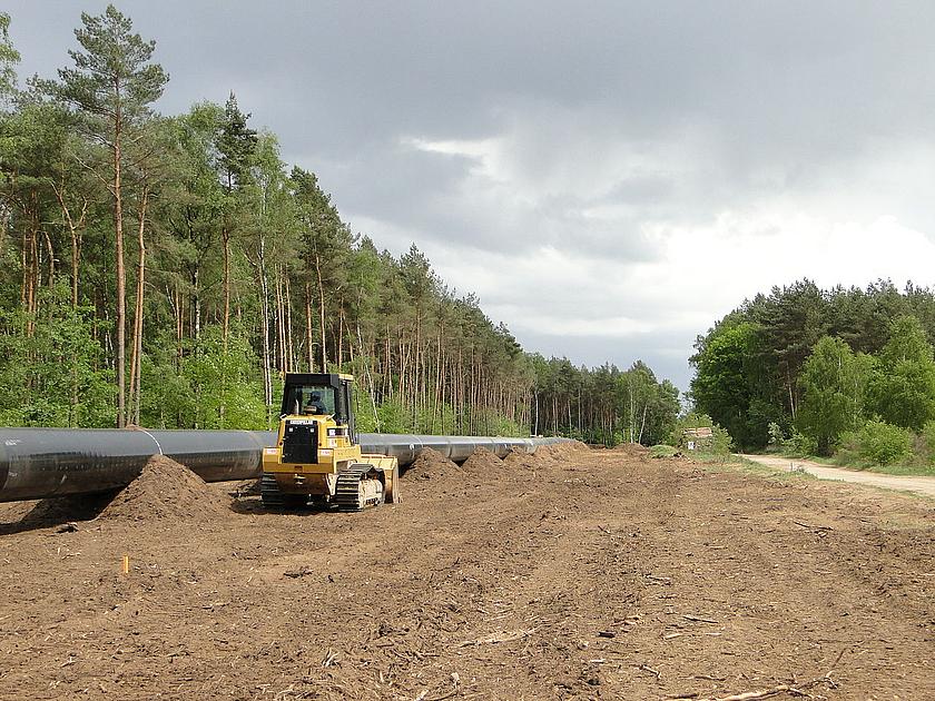 Bild einer im Bau befindlichen Erdgaspipeline. Im Hintergrund Wald und im Vordergrund Bagger und ein langes schwarzes Rohr.