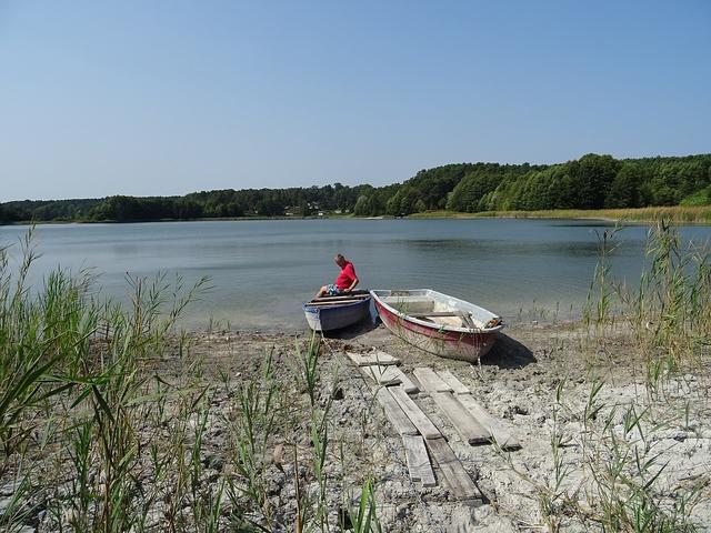 Ein Boot mit einem Mann am Ufer eines Sees dessen Üferböschung von einem sinkendem Wasserspiegel geprägt wirkt.