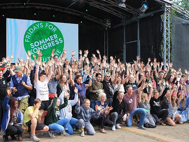 Gruppenbild vieler Menschen mit ausgestreckten Händen in den Himmel vor einer Bühne auf der ein Logo mit der Aufschrift Fridays for Future Sommerkongress prangt.