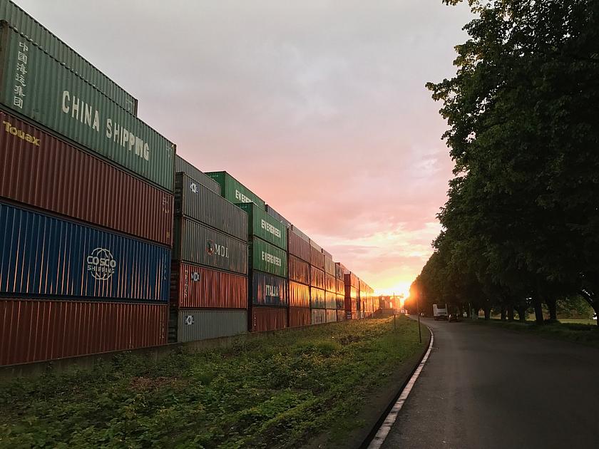 Links ragen gestapelte Container mit Waren auf. Rechts sind Bäume. In der Mitte eine Straße. Im Hintergrund der Sonnenuntergang.. 