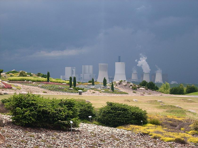Bild eines Parks mit Sträuchern und kleinen Bäumen, die beschriftet sind. Dahinter erheben sich die rauchenden Schornsteine eines Kohlekraftwerks.