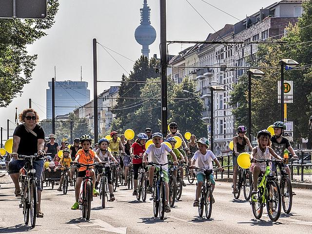 Eine Gruppe von Kindern mit gelben Ballons und Fahrrädern sind auf Berlins Straßen unterwegs.