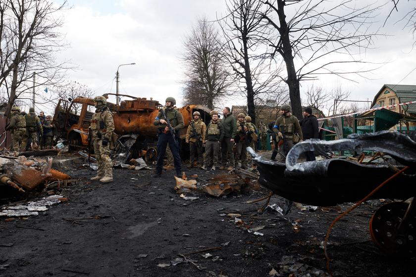 Männer und Frauen in schusssicheren Westen sowie in Soldatenmontur stehen auf einer zerstörten Straße voller Autowracks