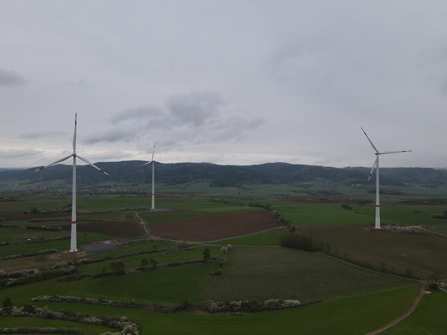 3 Windräder im bayerischen Franken vor einem Dorf und Bergen.