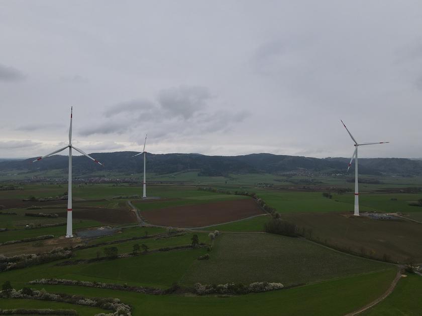 3 Windräder im bayerischen Franken vor einem Dorf und Bergen.