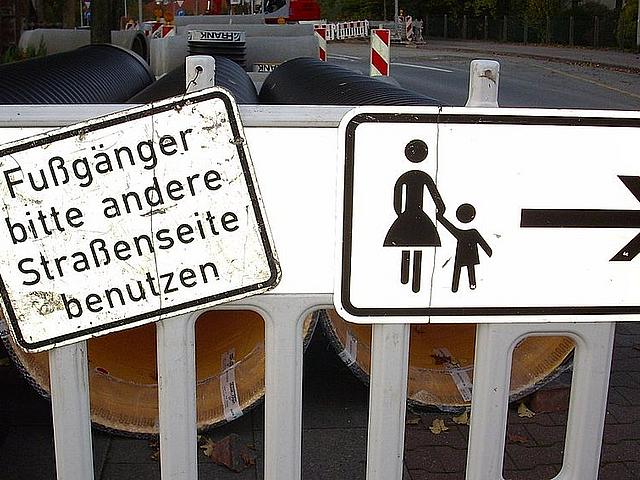 Straßenschild vor Baustelle auf dem Gehweg mit der Aufschrift: Fußgänger bitte andere Straßenseite benutzen