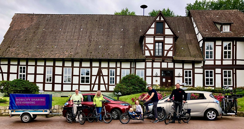Vier Erwachsene und zwei Kinder mit Autos und Fahrrädern vor einem Fachwerkhaus. 